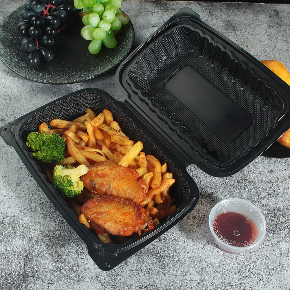 A black plastic clamshell container with an open lid containing French fries and chicken wings is placed on the table