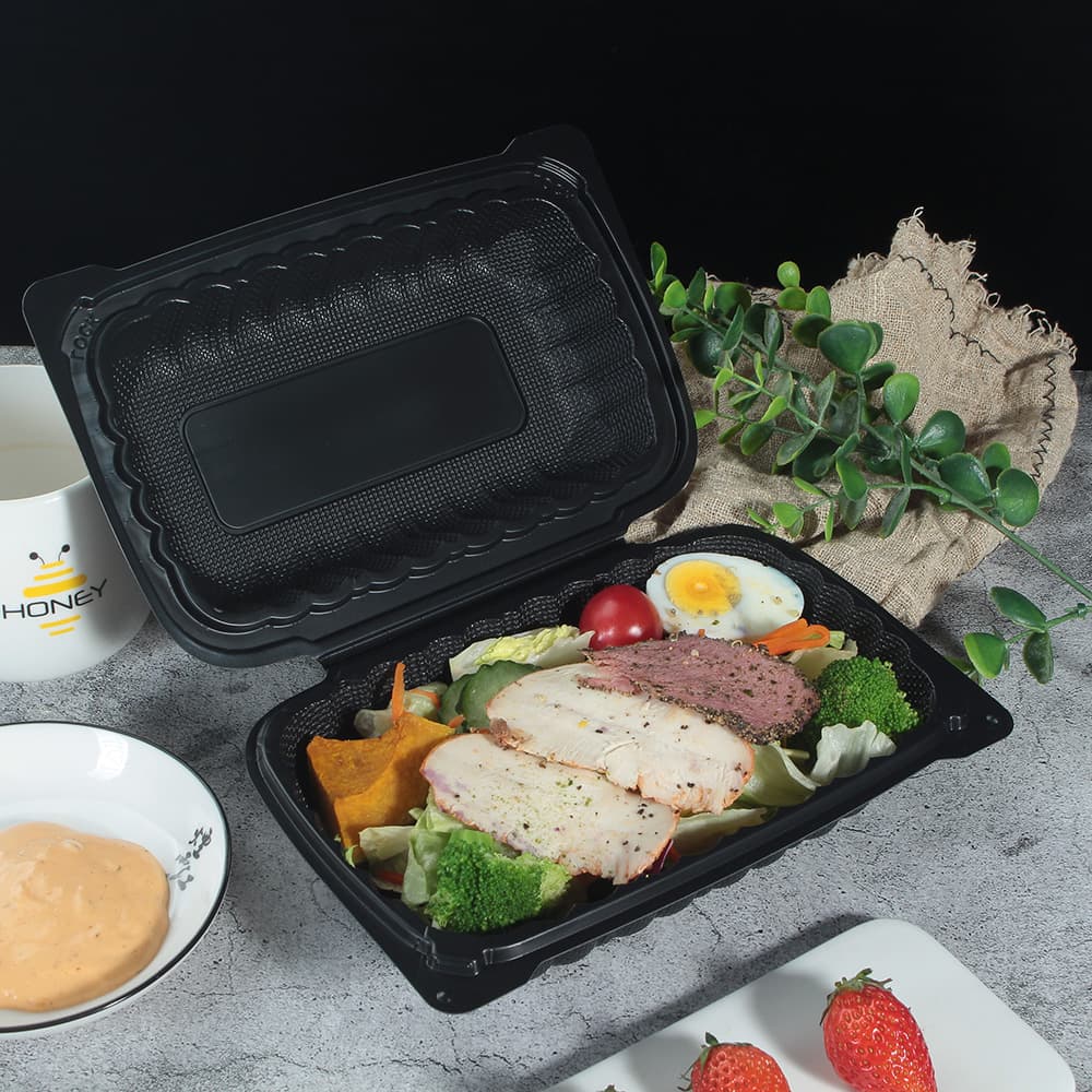 A black plastic clamshell container with the lid open to the right 45 degree view containing vegetables and meat slices is placed on the table