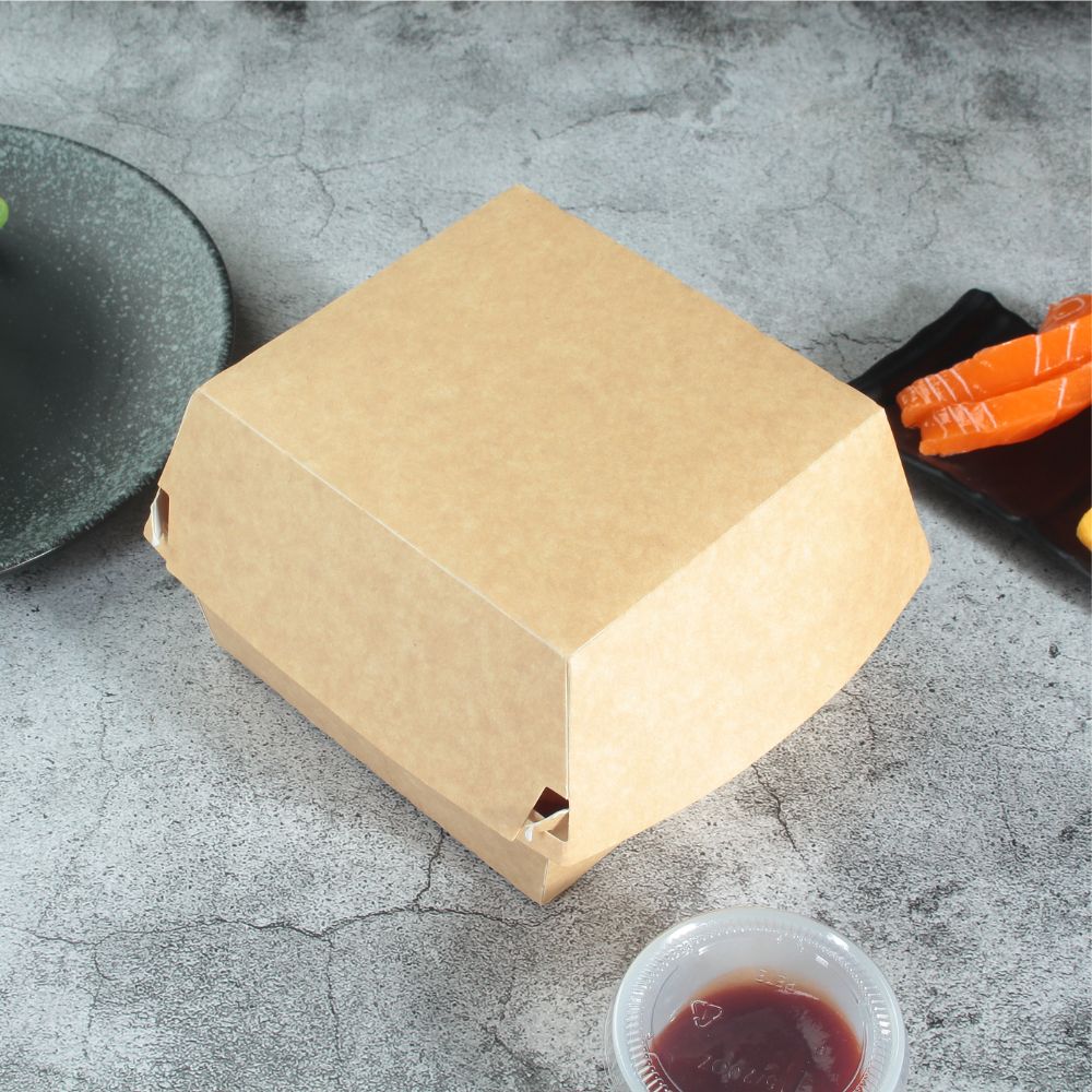 A closed kraft paper clamshell storage container lies on a gray table next to the edges of a saucer and red sauce in a clear portion cup