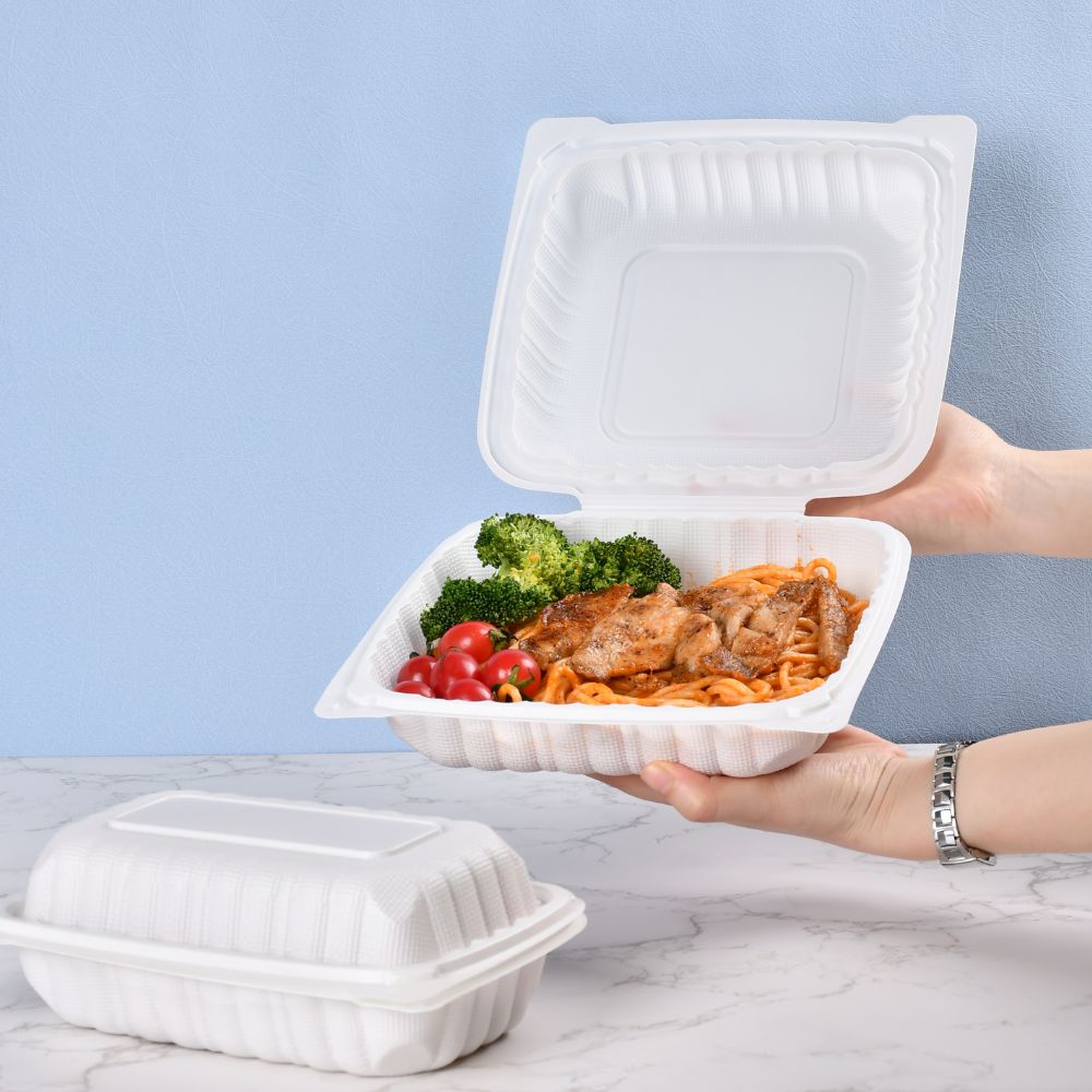 A rectangular clamshell bento box with the lid closed on the table and a clamshell bento box with a pair of hands holding the lid open and filled with pasta on a blue and white background.
