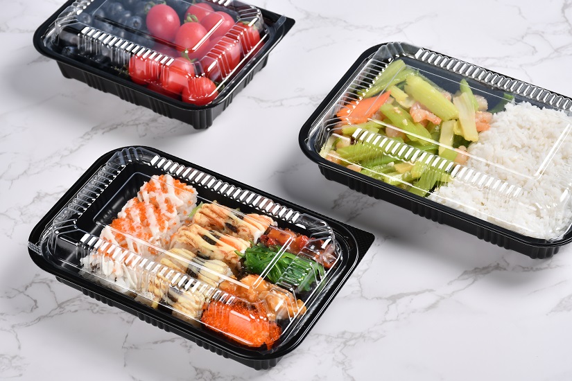 Three black lunch boxes of different sizes are placed on a gray background, containing fruits, sushi, rice and vegetables respectively.