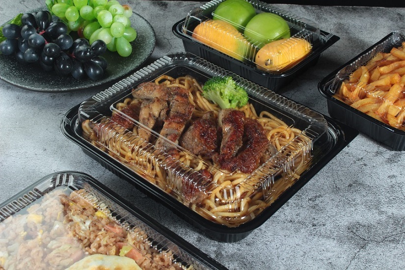 Four black rectangular plastic containers containing noodles, mangoes, rice and meat were placed on the table, decorated with green and purple grapes on round plates.