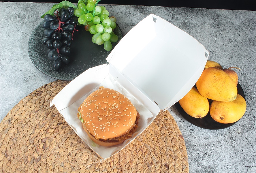 A kraft paper Clamshell box with the lid open, containing a burger, was placed on a table alongside green and purple grapes and mangoes on round plates.