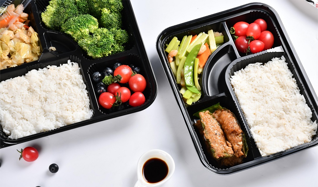 Two black 5-compartment lunch boxes without lids contain rice, vegetables and meat, tomatoes and blueberries on a gray background.