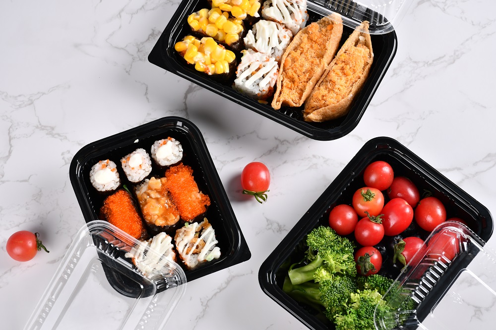 3 black plastic bento boxes are placed on an off-white table with two small tomatoes on it. Two bento boxes contain 8 pieces of sushi each, and one contains small tomatoes and broccoli with the transparent lid open and placed on the edge of the box.