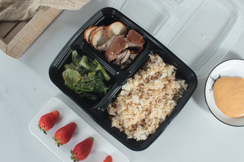 A three-compartment black lunch box filled with rice, meat and vegetables is placed on a white table. There is also a white rectangular plate with three strawberries and a bowl of yellow sauce on the table. A transparent lid is placed next to it, and the corner of a wooden box is exposed in the upper left corner.