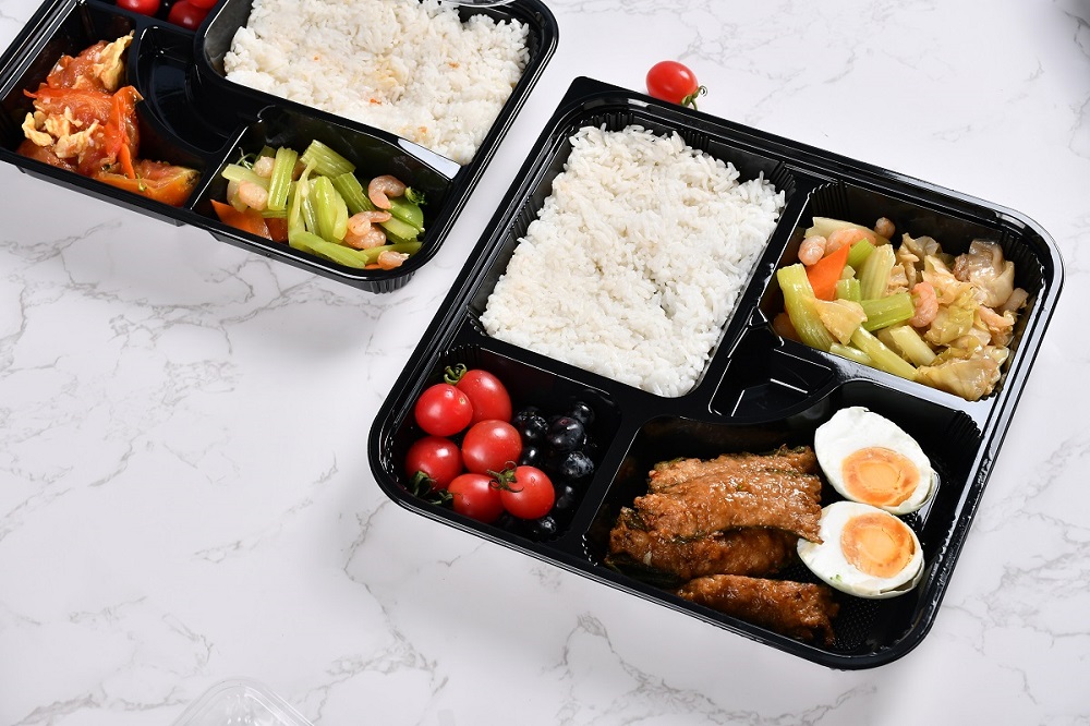 A 5-style lunch box containing celery, shrimp, green pepper wrapped in minced meat, eggs, cherry tomatoes, blueberries and rice, and a 5-grid lunch box also containing celery, shrimp and scrambled eggs with tomatoes are placed on a gray and white textured table.
