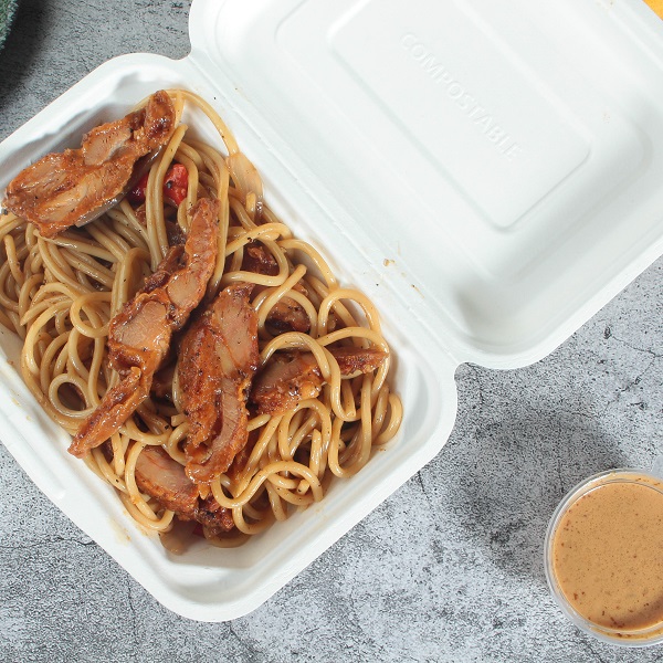 A zoomed-in white bagasse rectangular clamshell container with noodles and chicken opened and lying flat on a gray cracked table with a transparent portion cup of yellow dipping sauce on the table.