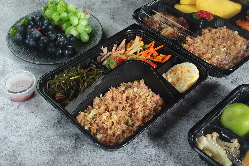 A black 5-grid plastic lunch box filled with ham fried rice, kelp shreds, corn and carrot salad and a fried egg was placed on an off-white table. There was also a plate on the table with a bunch of purple and a bunch of green grapes.