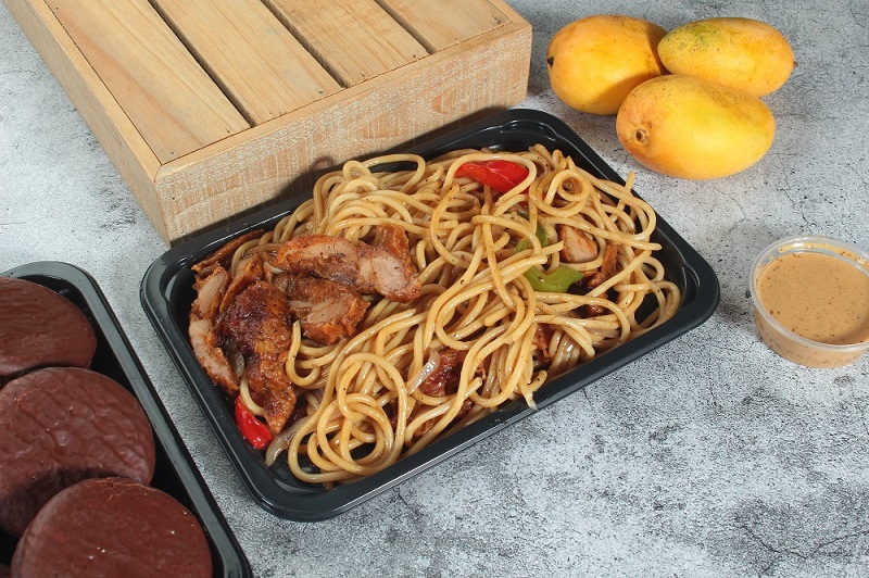 A black bento box containing pasta and chicken sits on a grey textured table with a wooden board, 3 mangoes, a bowl of hummus in a clear sauce cup, and part of another black bento box containing 3 chocolate chip cookies.