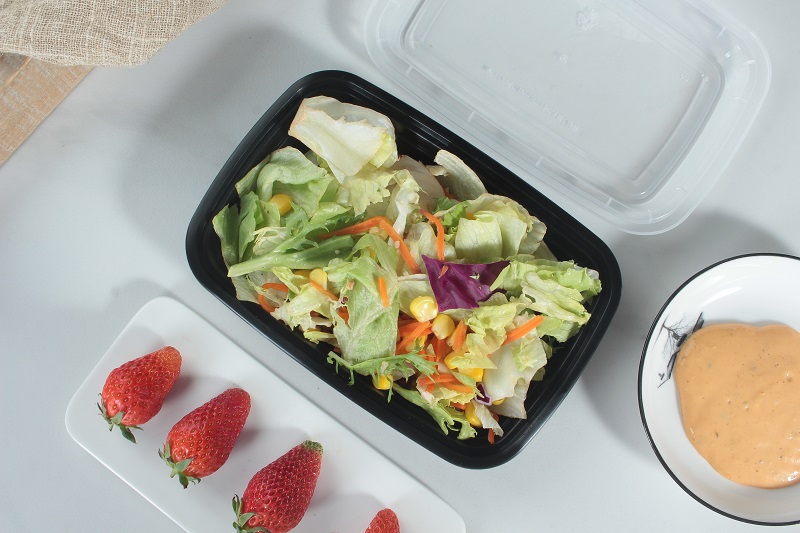 A black plastic bento box filled with various vegetables, carrots, corn and cucumber is placed on a white table. There is also a white rectangular plate with 3 strawberries and a bowl of yellow sauce on the table. The transparent lid is also placed next to it.