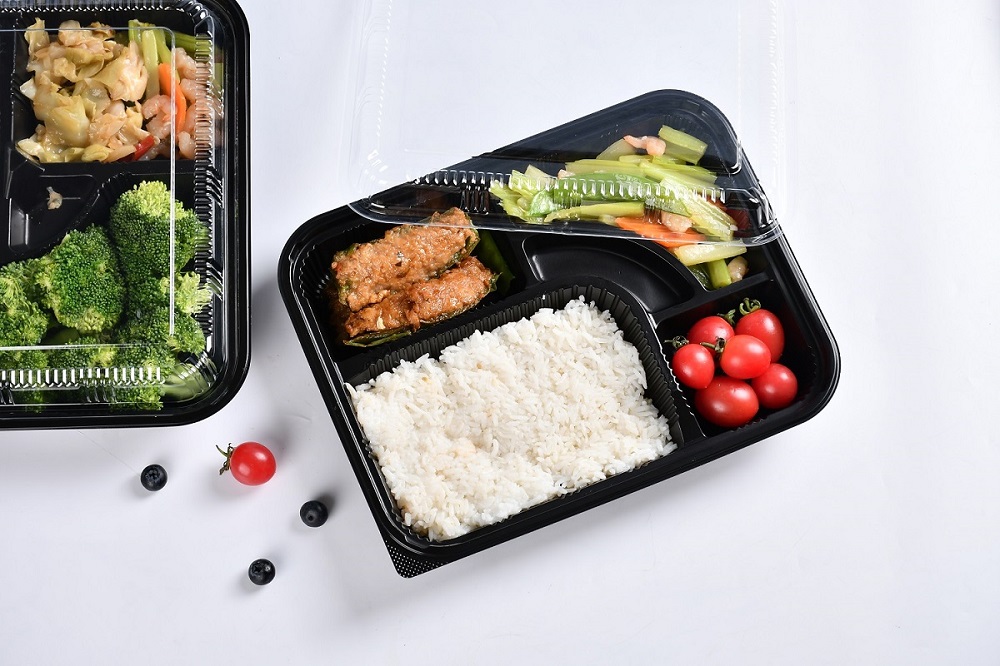 A black 5-grid plastic bento box was placed on a gray-white table with two blueberries and a small tomato scattered on it. Inside the bento box were rice, meat patties, small tomatoes, blueberries, celery and carrots. There was another bento box with only a small part exposed and it also contained broccoli, celery and shrimp.