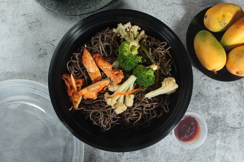 A black round plastic bowl filled with soba noodles, chicken and broccoli is placed on a grey textured table with a plate containing 3 mangoes and a cup of red sauce next to it.