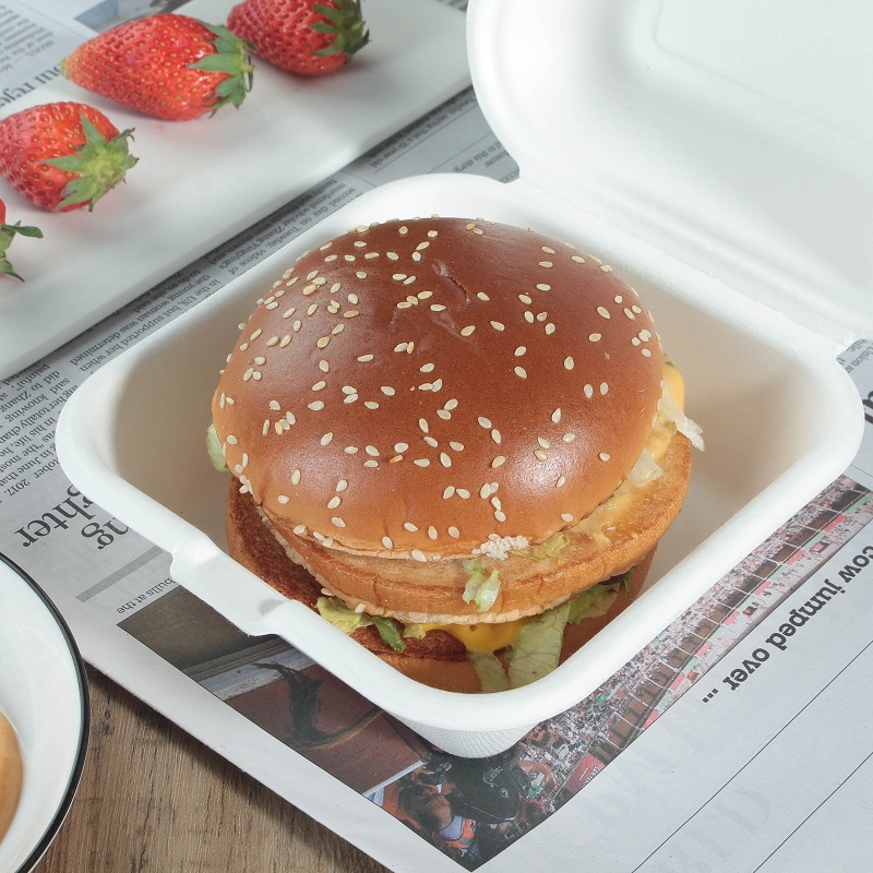 A clamshell container with a burger inside, zoomed in on a tabletop covered with newspapers and some strawberries