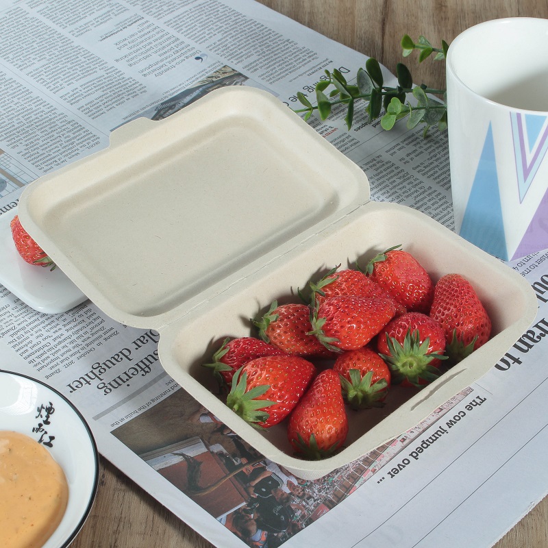 A natural color bagasse rectangular clamshell container is filled with red strawberries and placed on a table covered with newspaper and decorated with cups, leaves and a bowl of yellow sauce in a round plate