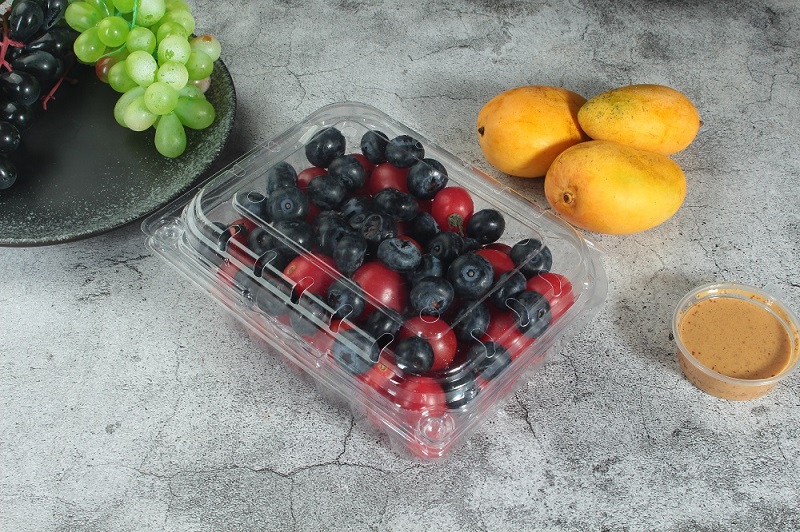 A clear perforated clamshell container with cherry tomatoes and blueberries is placed on an off-white table. There is also a gray plate with green and purple grapes, 3 mangoes and a cup of yellow sauce on the table.