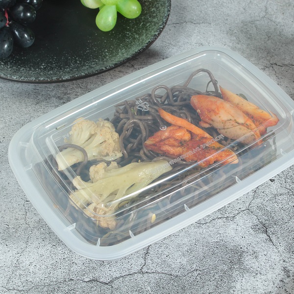 A transparent disposable plastic bento box with black noodles, meat, and vegetables is placed on a gray table decorated with fruits