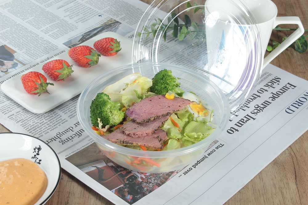 A transparent plastic round bowl filled with vegetables, carrots, corn and meat slices was placed on a table covered with newspapers. On the table there was a cup, a white rectangular plate with 4 strawberries, a bowl of yellow sauce and a leaf.