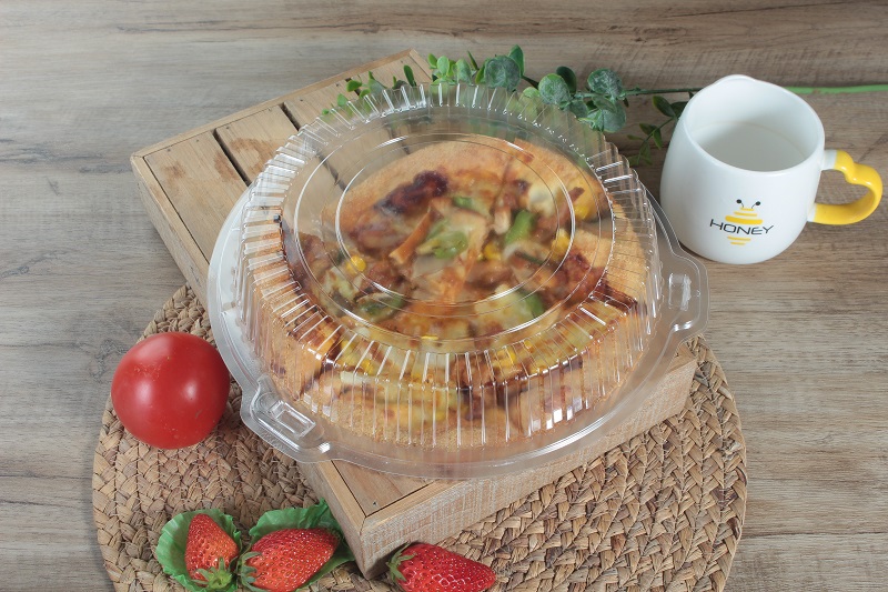 A transparent round clamshell bento box contains a round cheese chicken pizza and is placed on a straw mat on a table with a tomato, three strawberries, a white and yellow cup and a leaf on top.