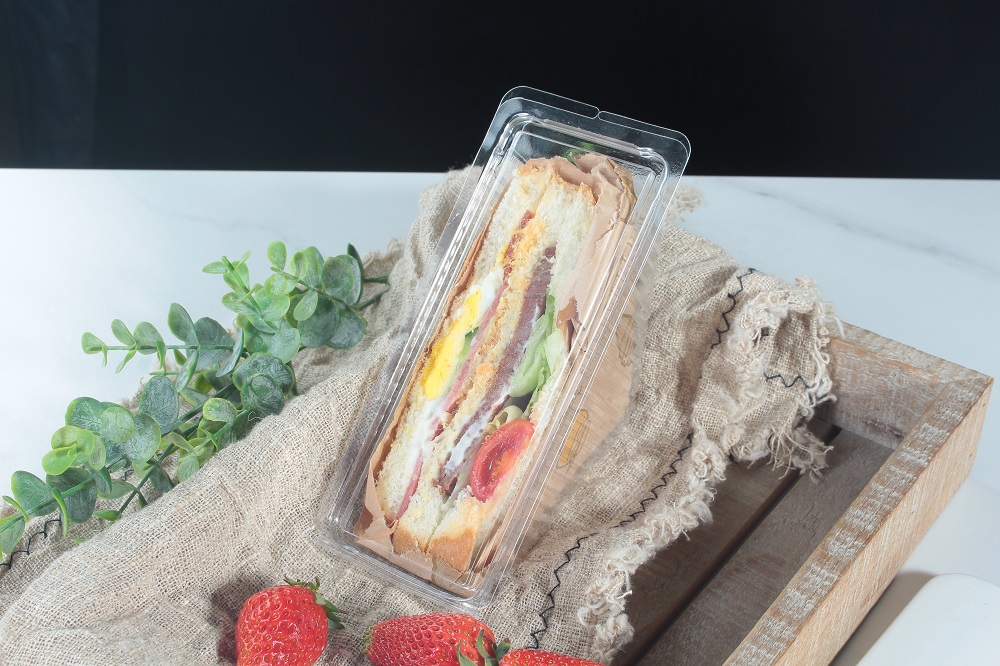 A ham, egg and pork floss sandwich is placed in a transparent triangular container and placed in a wooden box. Inside the box there is a piece of linen, 3 strawberries and a leaf next to them.