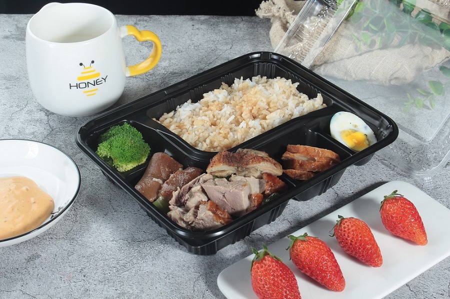 A black 5-grid bento box filled with grilled chicken, eggs, broccoli and rice is placed on a gray-white table. There is also a cup, a white rectangular plate with 4 strawberries, a bowl of yellow sauce, a leaf and a linen strip on the table, with the lid open and placed next to it.