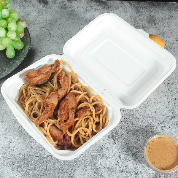 A white bagasse rectangular clamshell container filled with noodles and chicken and opened flat on a gray cracked table with green grapes and a transparent portion cup with yellow dipping sauce.