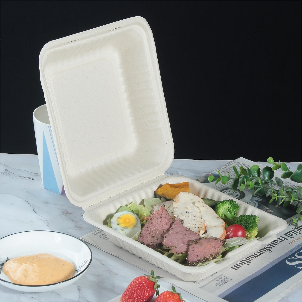 Detail of a 3-compartment compostable bagasse clamshell box filled with vegetable and meat slices placed on a tabletop covered with newspaper and a bowl of yellow sauce, leaves, and strawberries on a black background.