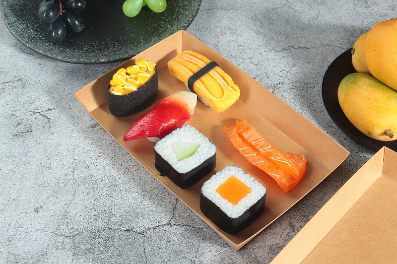 A rectangular kraft paper sushi tray holds five pieces of sushi and two pieces of salmon, and is placed on an off-white table with purple and green grapes and mangoes.