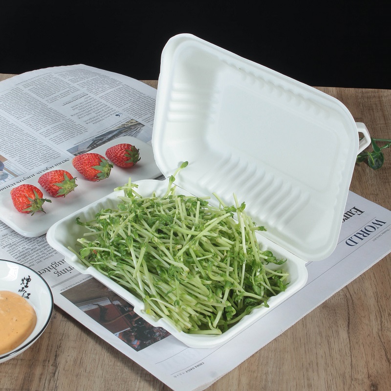 A white bagasse rectangular clamshell container filled with green vegetables is placed on a wooden table covered with newspaper, a bowl of yellow sauce and strawberry decoration