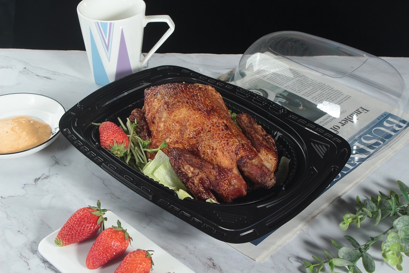 A black roast chicken box containing a roast chicken, a strawberry and some vegetables was placed on a gray-white table covered with newspapers. There was also a cup, a white rectangular plate with 3 strawberries, a bowl of yellow sauce, and a leaf on the table with the lid open.