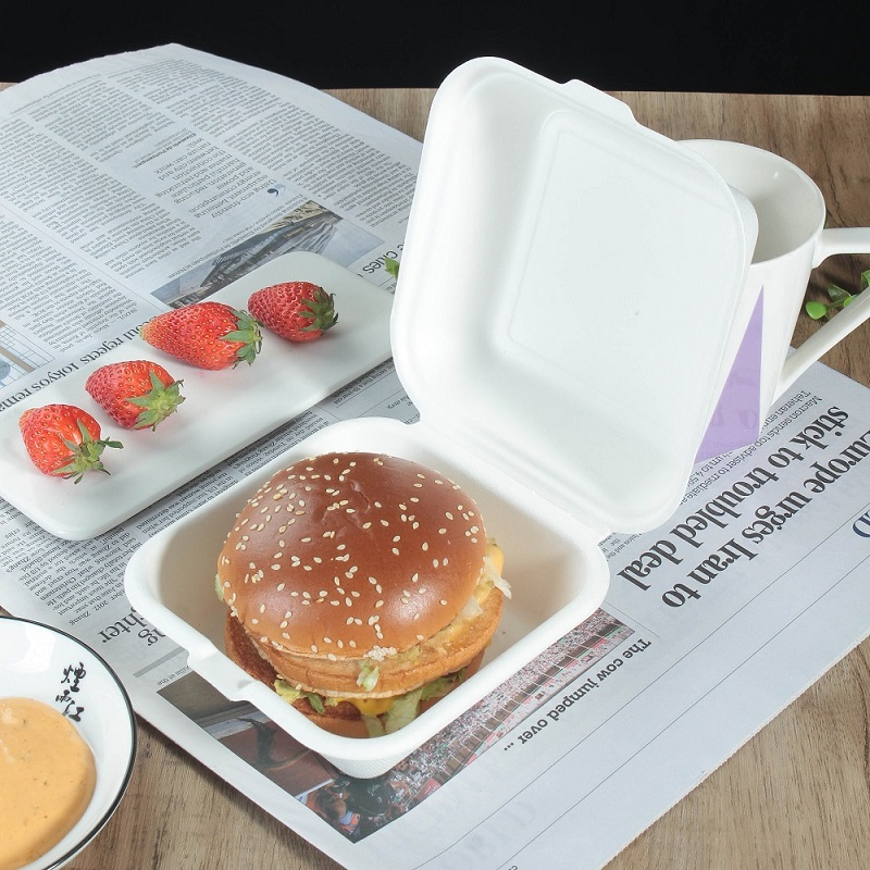 A clamshell container with hamburgers in it, opened and lying on a table covered with newspapers, with some strawberries and a bowl of yellow sauce on the table