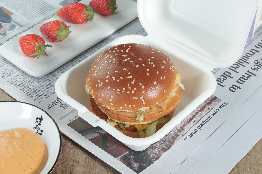 A white bagasse clamshel bento box with a cheese beef burger is placed on a wooden table covered with newspapers, and there are 4 strawberries on a rectangular plate and a dish of yellow sauce on the wooden table.