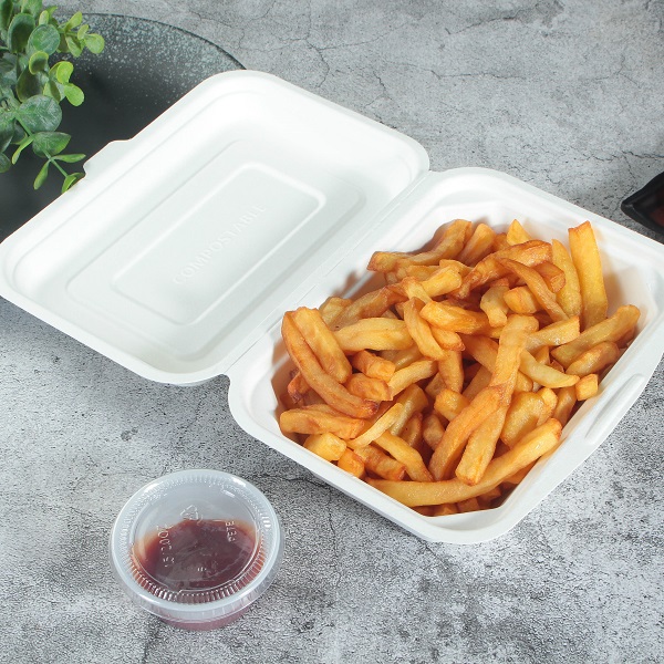 A white bagasse rectangular clamshell container filled with French fries and opened flat on a gray cracked table with leaves and a transparent split cup with red dip.